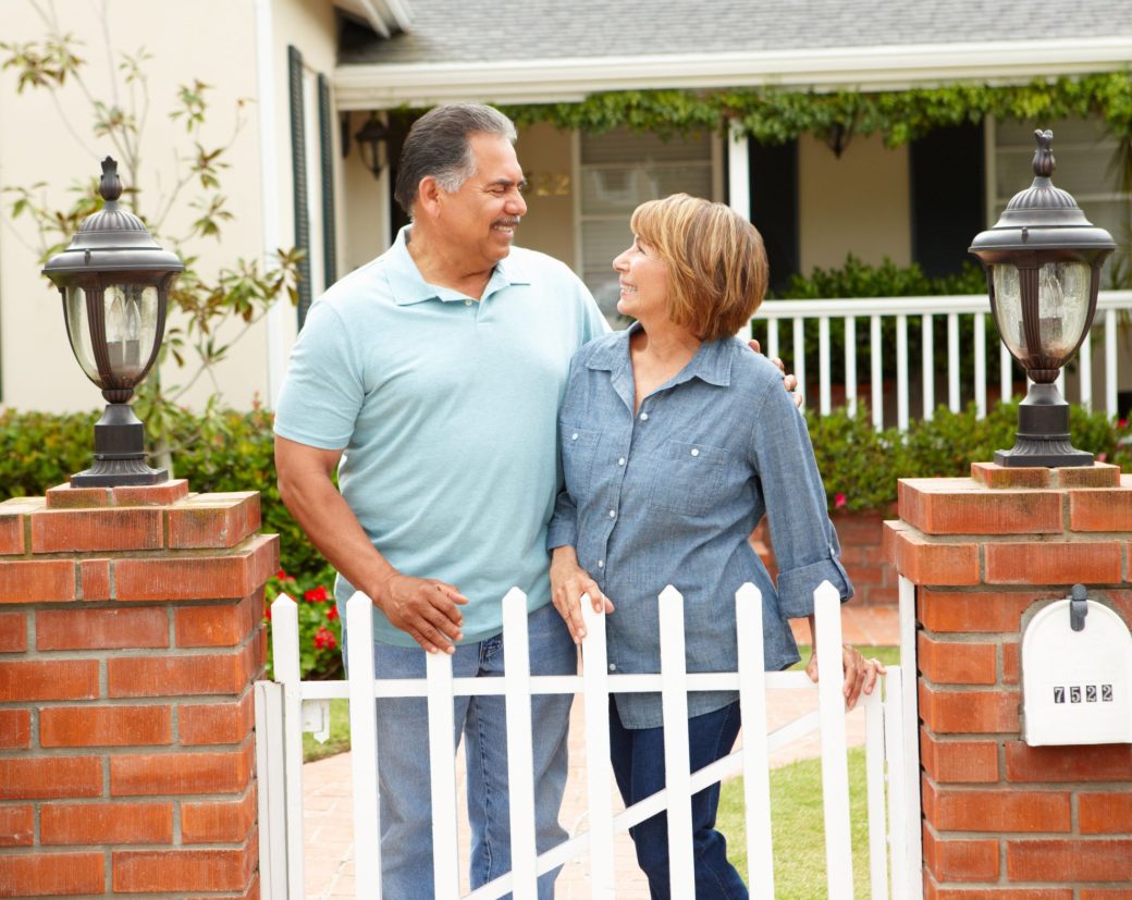 An older couple smiling at each other, standing by a white picket gate.
