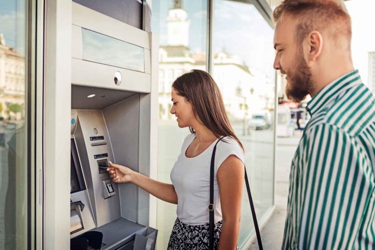 A person inserts a card into an ATM as another person in a striped shirt stands nearby.