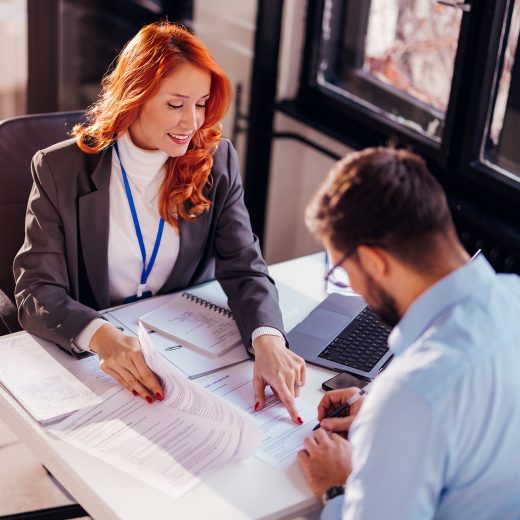 A woman points to a document while a man signs it at a desk with a laptop.