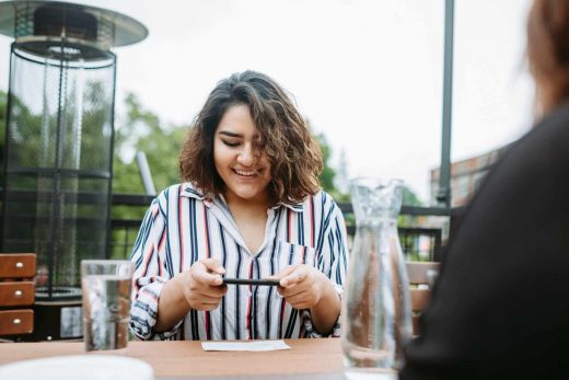 A smiling woman takes a photo of a check on a table with phone at an outdoor cafe.