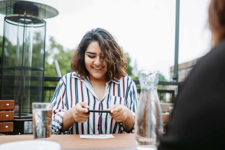 A smiling woman takes a photo of a check on a table with phone at an outdoor cafe.