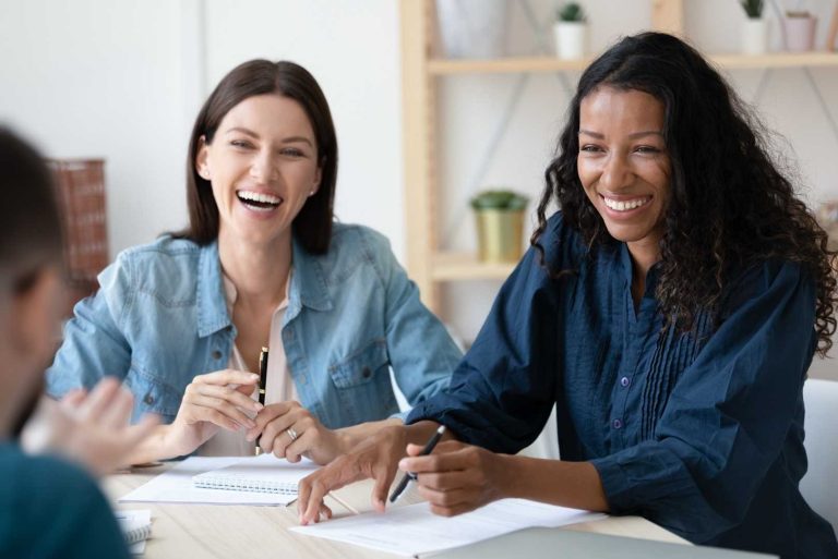 Two people are laughing while sitting at a desk with papers and pens.
