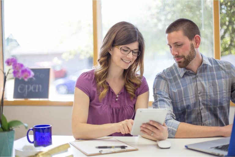 A woman wearing glasses and a purple shirt looks at a tablet with a man in a blue plaid shirt.
