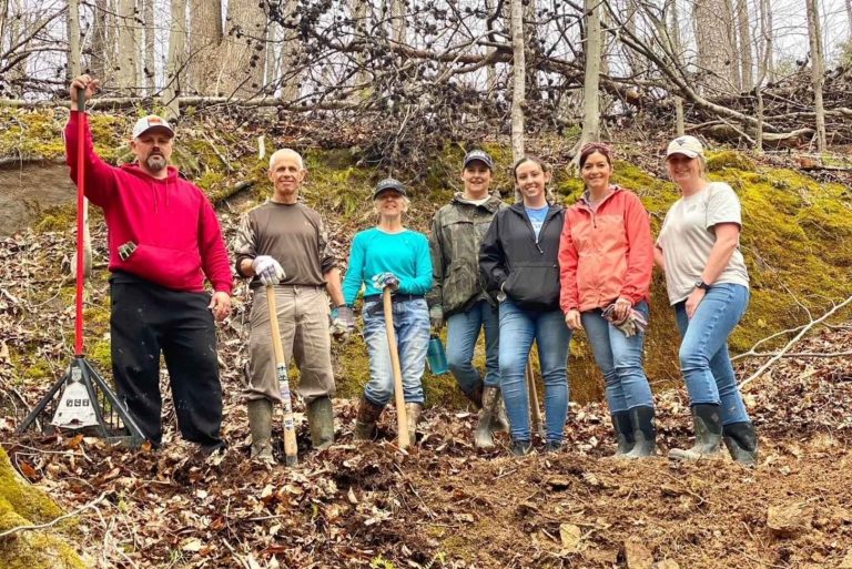 A group of people stand in a wooded area with yard tools