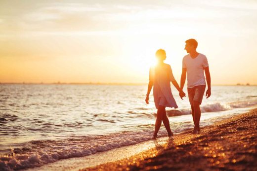 Silhouettes of a couple holding hands while walking on a beach at sunset.
