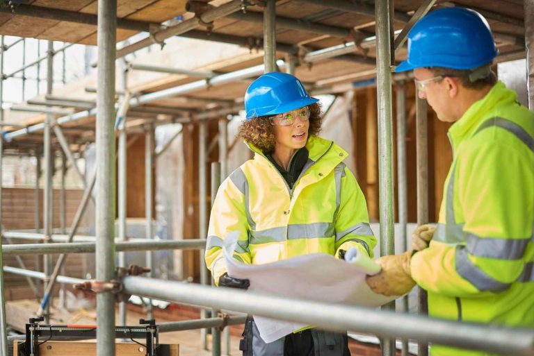 Two construction workers in hard hats and high-visibility jackets look at blueprints on a construction site.