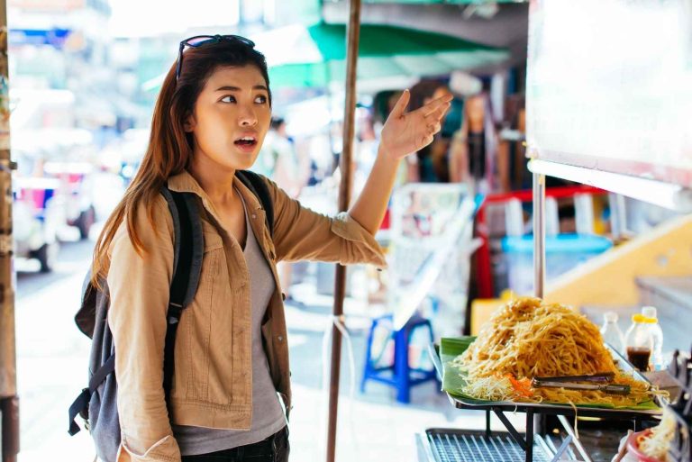 A person stands in front of a street food stall, gesturing towards a large mound of noodles.