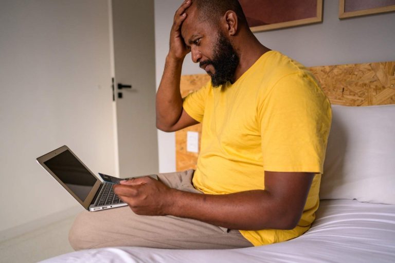 A person sits on a bed with a laptop and credit card, holding their head in distress.