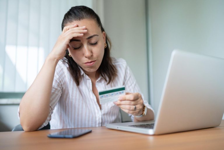 A person holding a credit card and looking stressed while sitting in front of a laptop and phone.