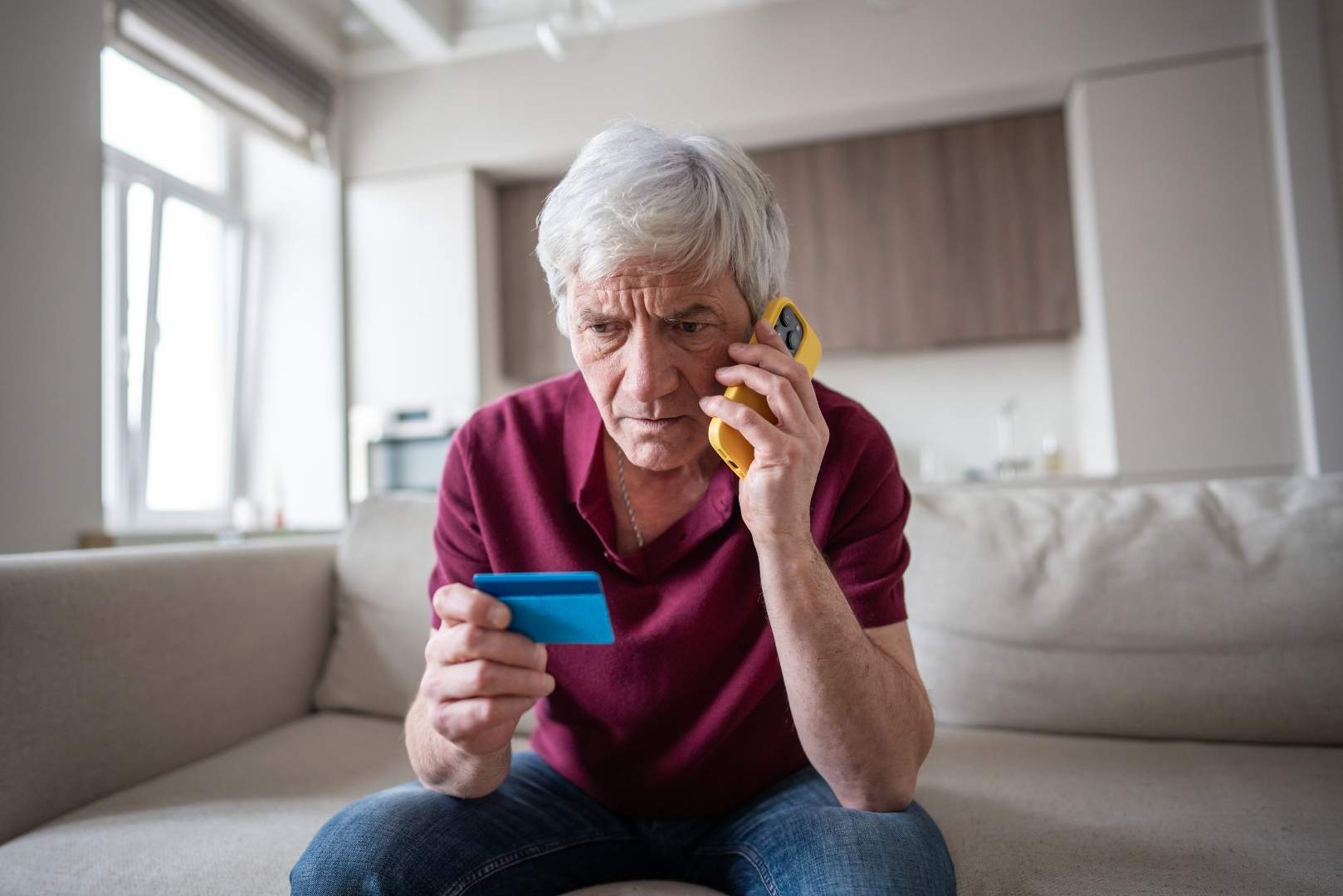 A man with gray hair is sitting on a couch, talkin on the phone while holding a credit card.