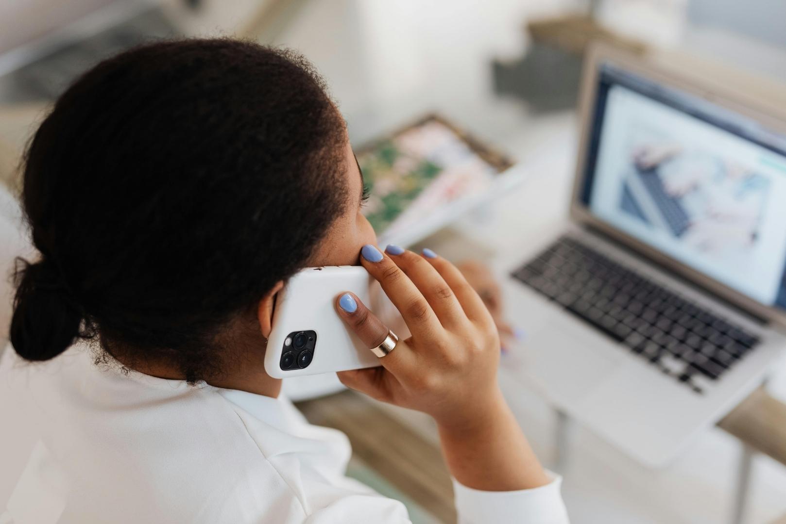 A person is talking on a white smartphone, with a laptop open in front of them.