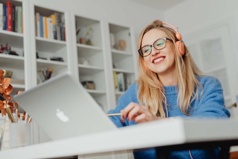 A woman with glasses and headphones smiles while looking at a laptop in front of a bookshelf.