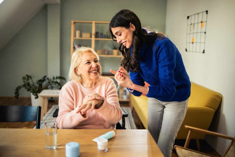 A caregiver an older woman apply lotion to her hands while they sit at a table.