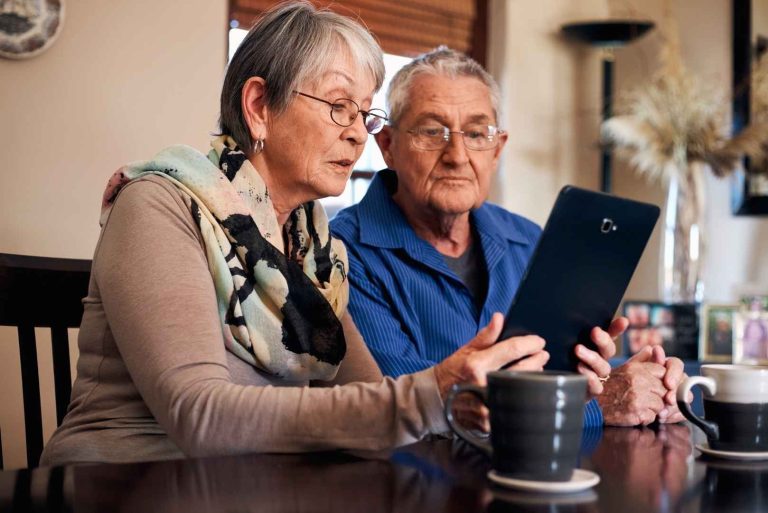Two older people looking at a tablet together while sitting at a table with mugs of coffee.
