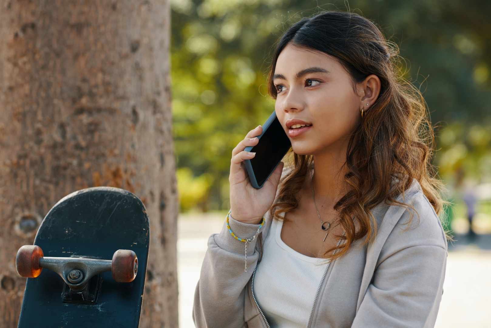 A young girl is talking on a mobile phone next to a skateboard and a tree outdoors.