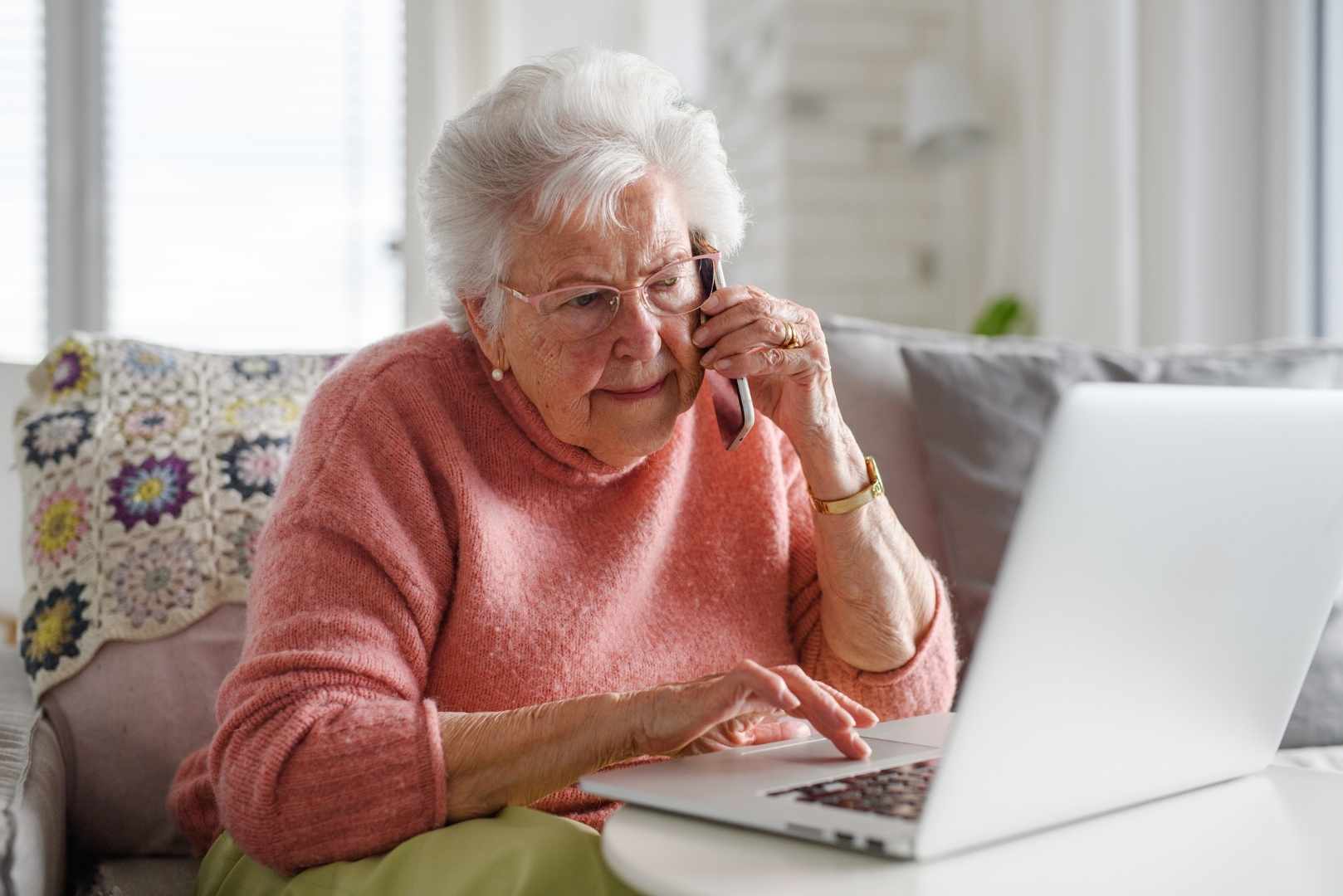 An older woman talks on a phone while typing on a laptop.