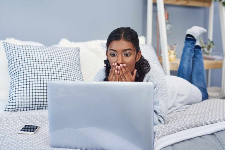 A person lies on a bed, looking at a laptop with a shocked expression, hands covering their mouth.
