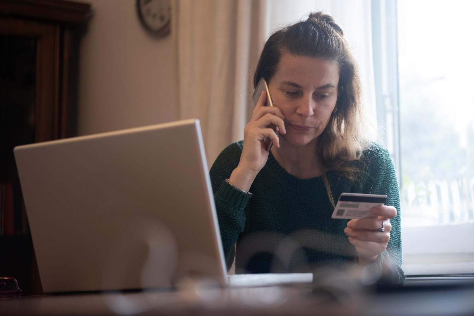 A woman on the phone holding a credit card in front of an open laptop.