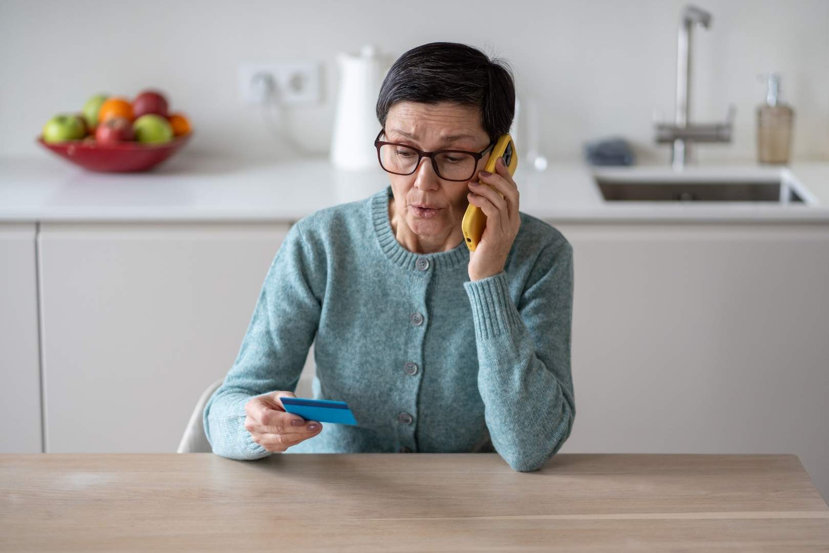 An older person is sitting at a kitchen table, holding a credit card and talking on a phone.