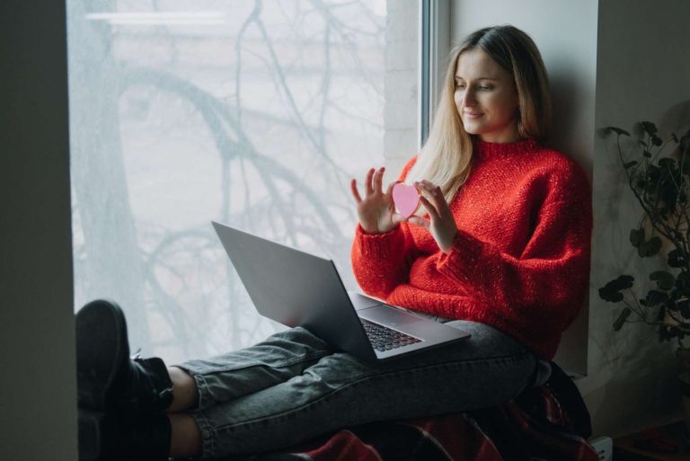 A person wearing a red sweater sits by a window with a laptop and holds a pink heart-shaped object.
