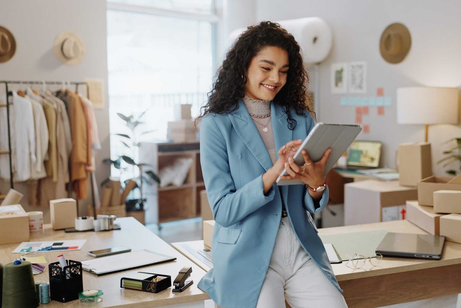 A woman smiles while looking at a tablet in a room filled with clothing racks, boxes, and supplies.