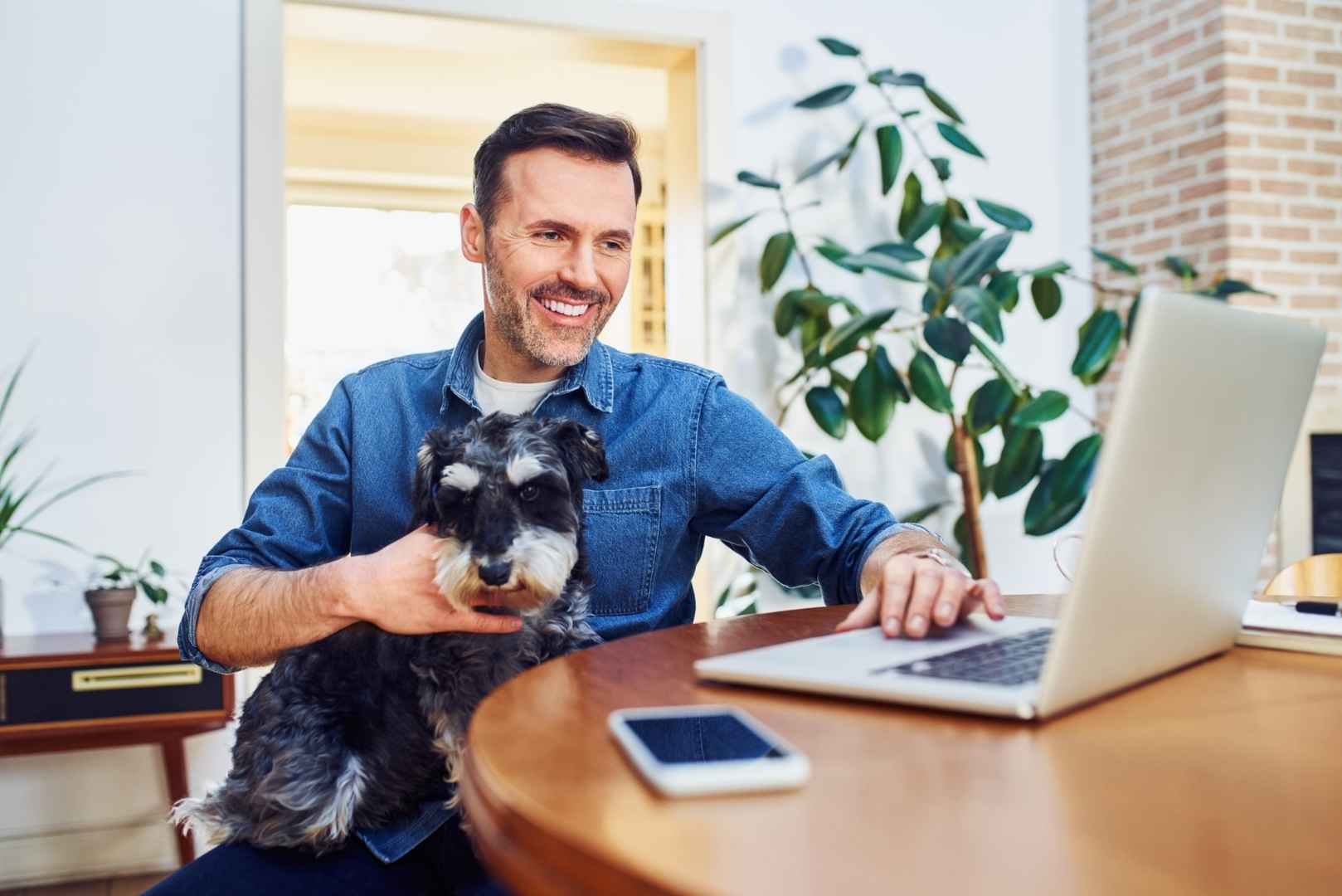 A man holds a small dog while working on a laptop at a wooden table.