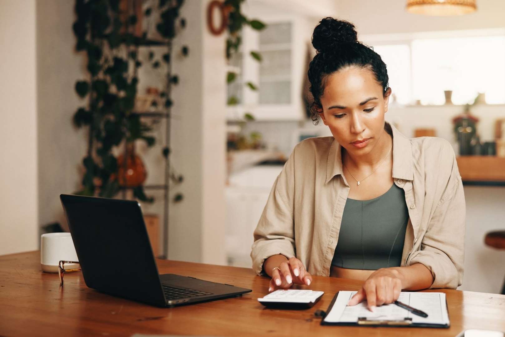 A woman sits at a table with a laptop, calculator, and papers, appearing to be working on finances.