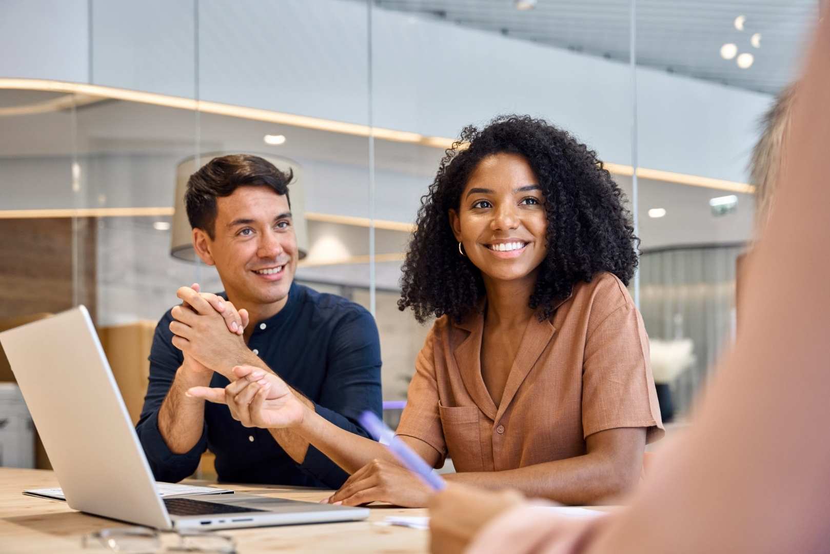 A man and a woman are sitting at a table with a laptop and looking at a person while smiling.