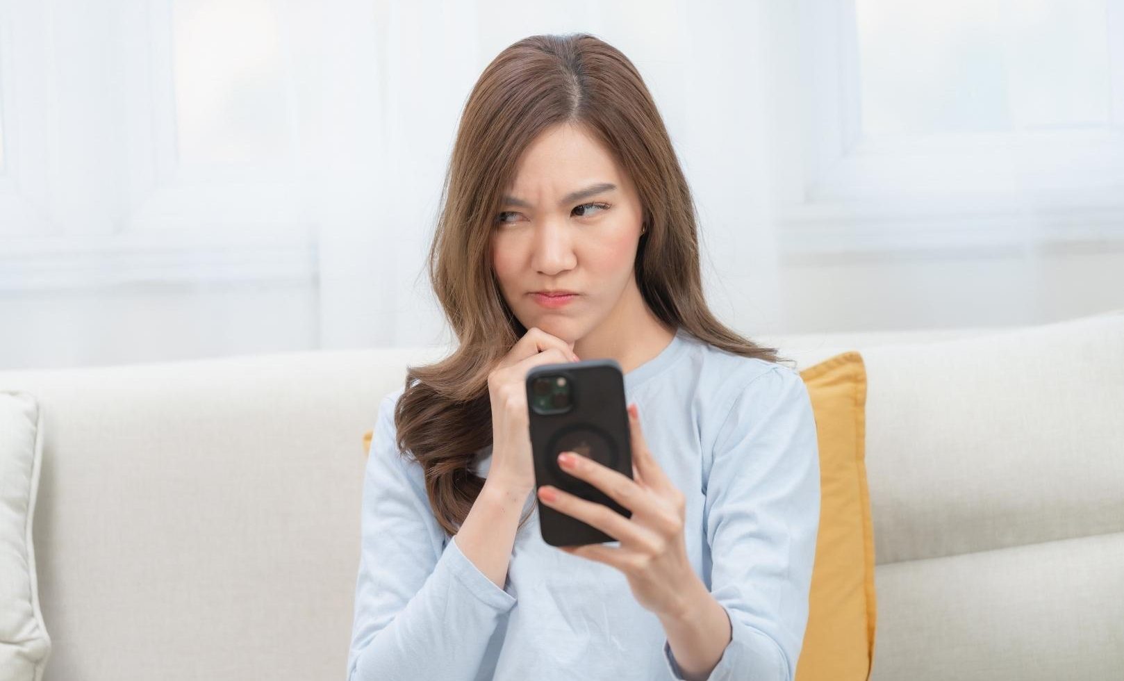 A woman is sitting on a couch and looking at their phone with a confused expression.