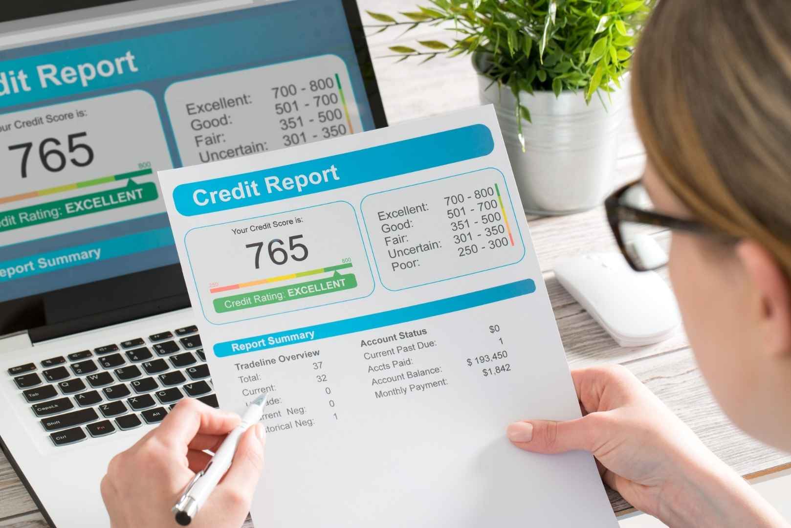 A person reviews a credit report showing a score of 765 with a laptop displaying similar information in the background.