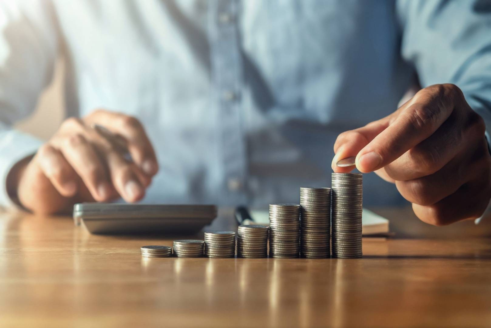 A person stacking coins on a wooden desk, with a calculator and notebook in the background.