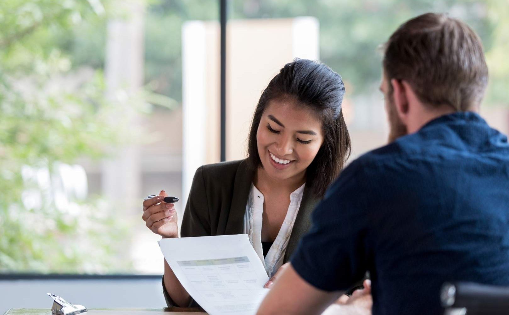 A woman smiles while holding a pen and looking at a document with a man seated opposite her.