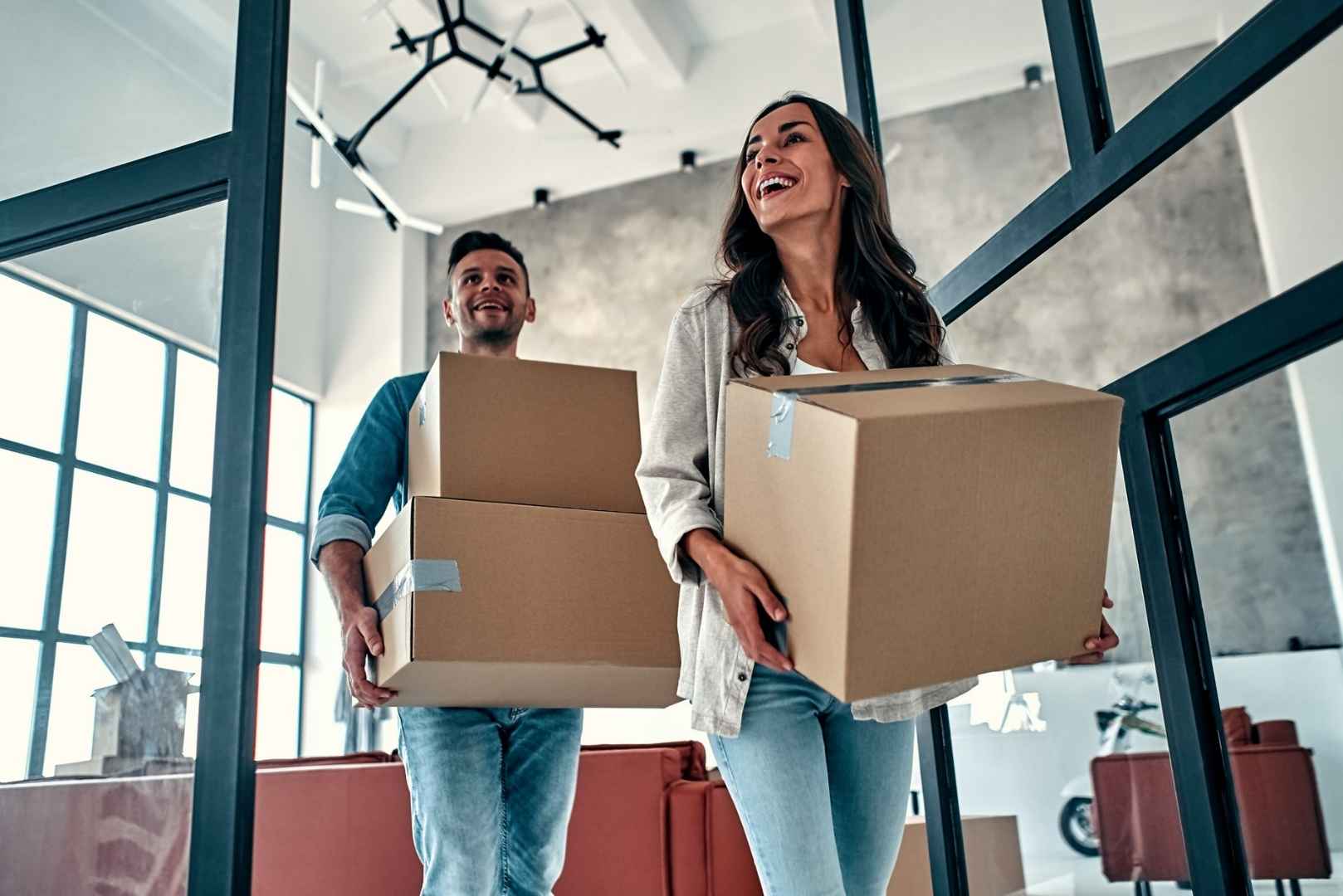 A smiling couple carries cardboard boxes into a new home.