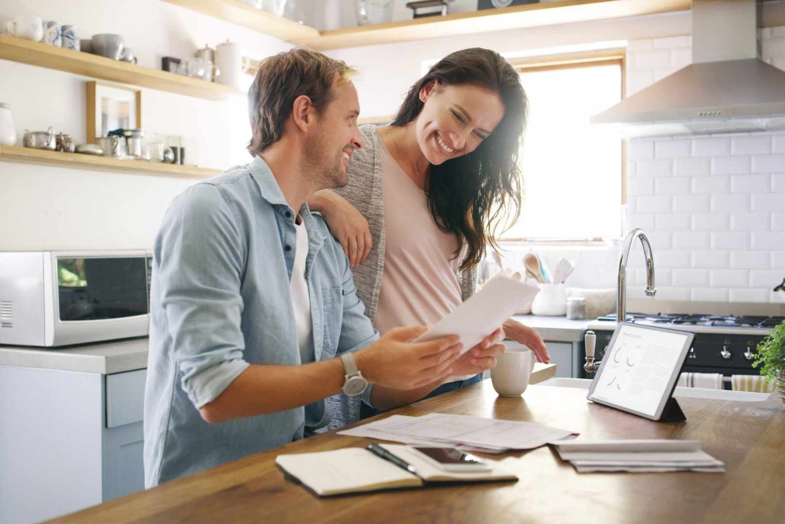 A couple smiles at each other in a kitchen while reviewing documents, with a tablet on the counter.