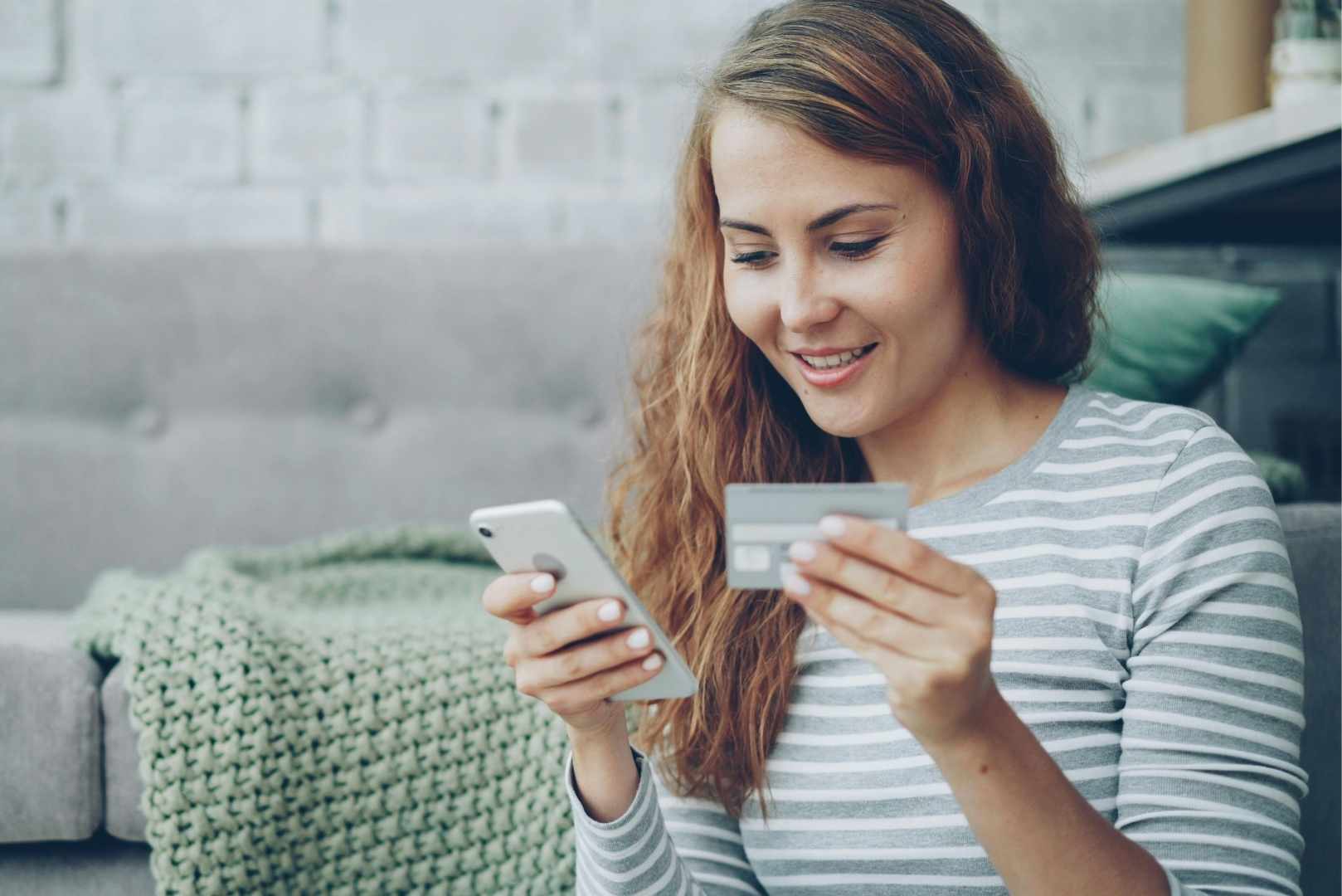 A woman smiles while holding a smartphone in one hand and a credit card in the other.