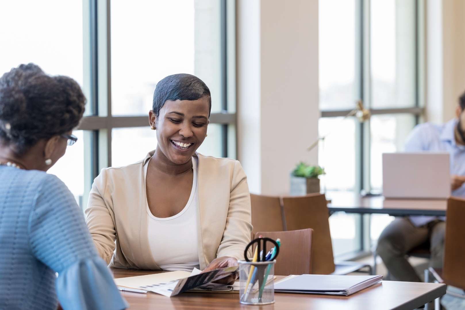 A smiling person looks down at papers on a table, with another person seated opposite them in an office space.