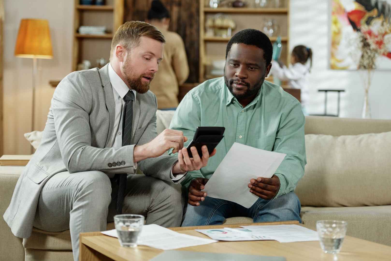 A man in a suit and tie uses a calculator while sitting next to another man holding papers on a couch.