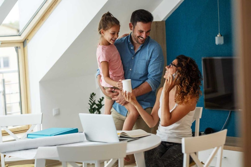 A smiling family interacts in a bright room while the mom talks on the phone at a table with a laptop.