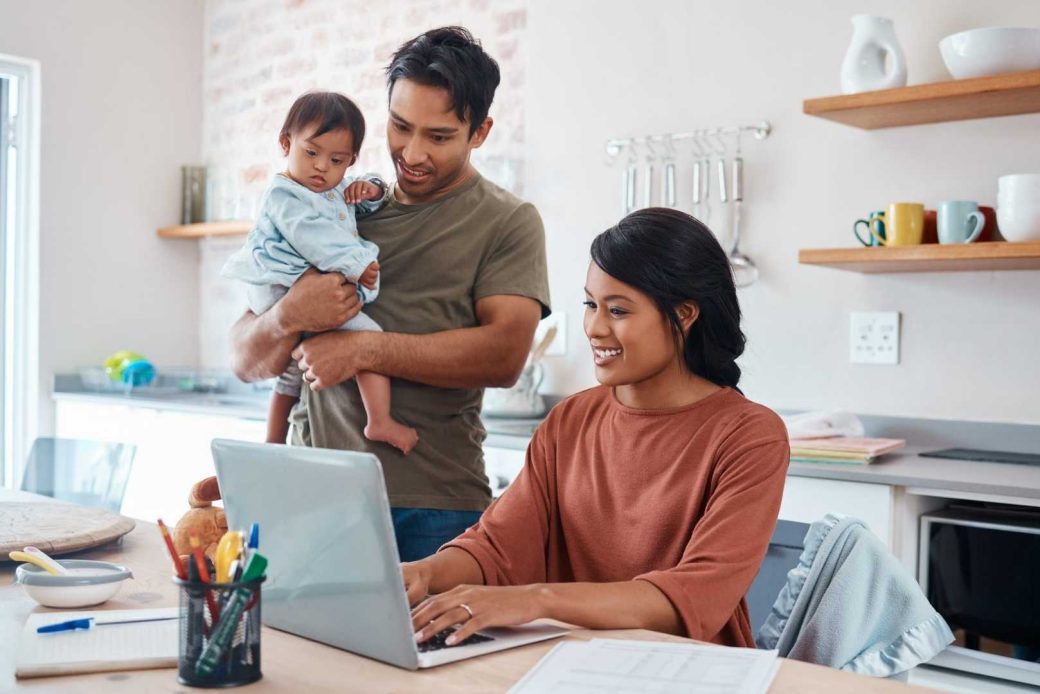 A woman works on a laptop while a man holds a baby standing behind her in a kitchen.