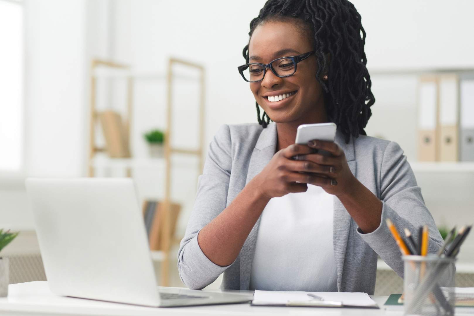 A woman wearing glasses smiles while holding her phone and looking at her laptop in front of her on a desk.