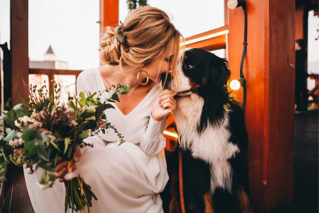 A bride in a white dress holds a bouquet of flowers while interacting with her dog.