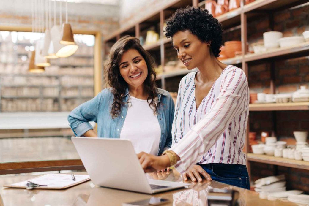 Two women in a pottery shop are looking at a laptop screen, one is pointing at the screen.