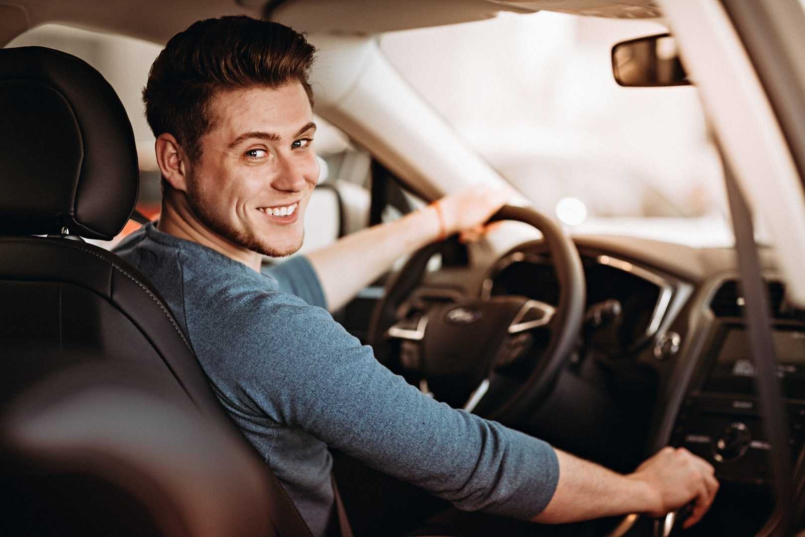 A smiling man sits in the driver's seat of a car, holding the steering wheel and looking over his shoulder.