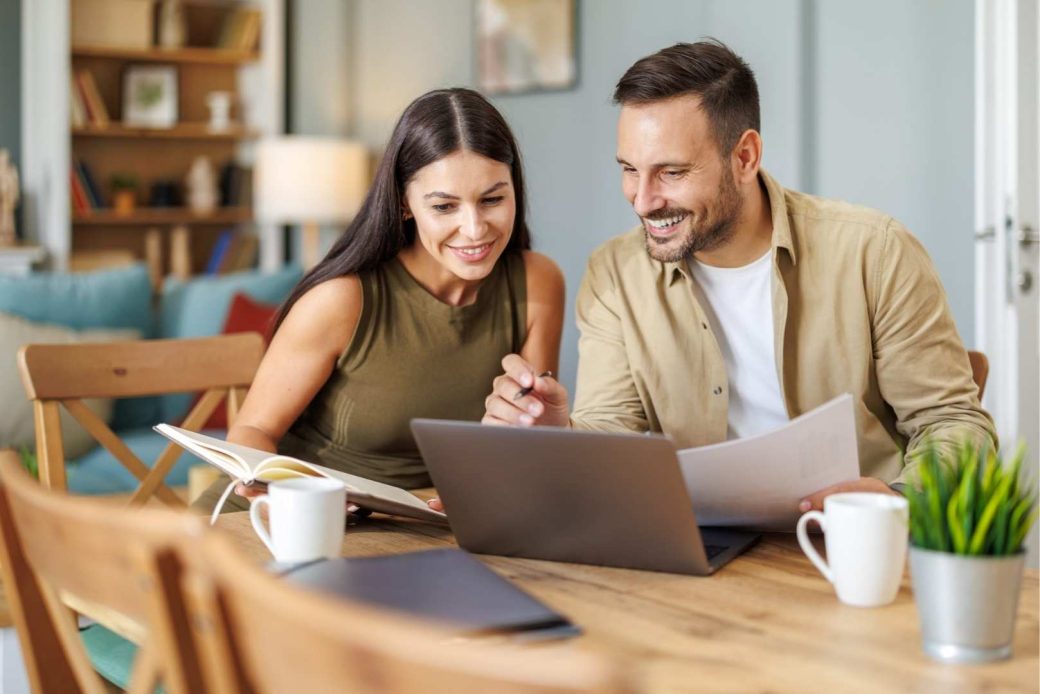 A man and a woman smiling while looking at a laptop and papers on a wooden table.