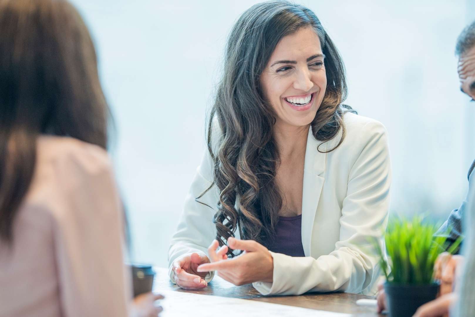 A woman in a white blazer laughs while talking to colleagues at a table.